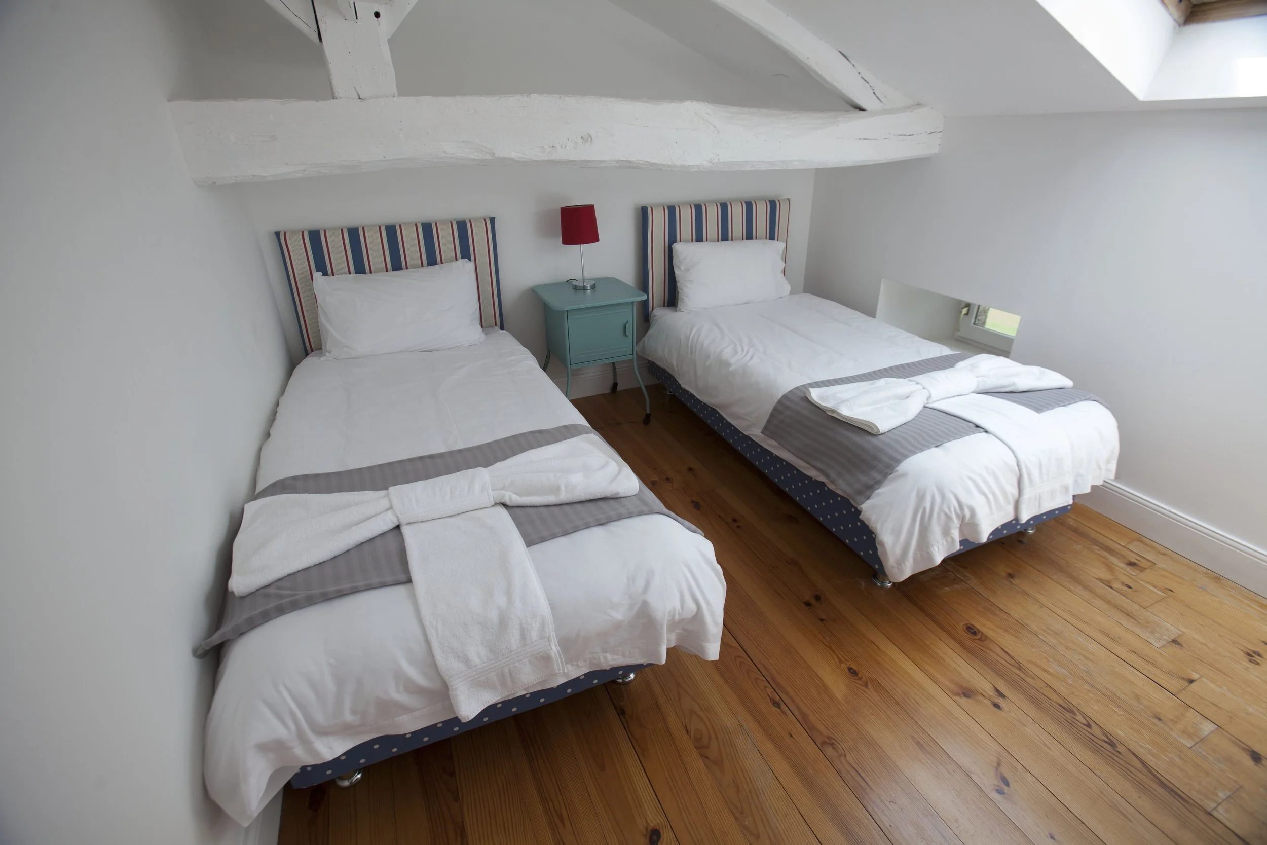 A bedroom with two twin beds with bedspreads and pillows, a small turquoise nightstand, and a red lamp. The room has white walls, a sloped ceiling with exposed beams, and a hardwood floor.