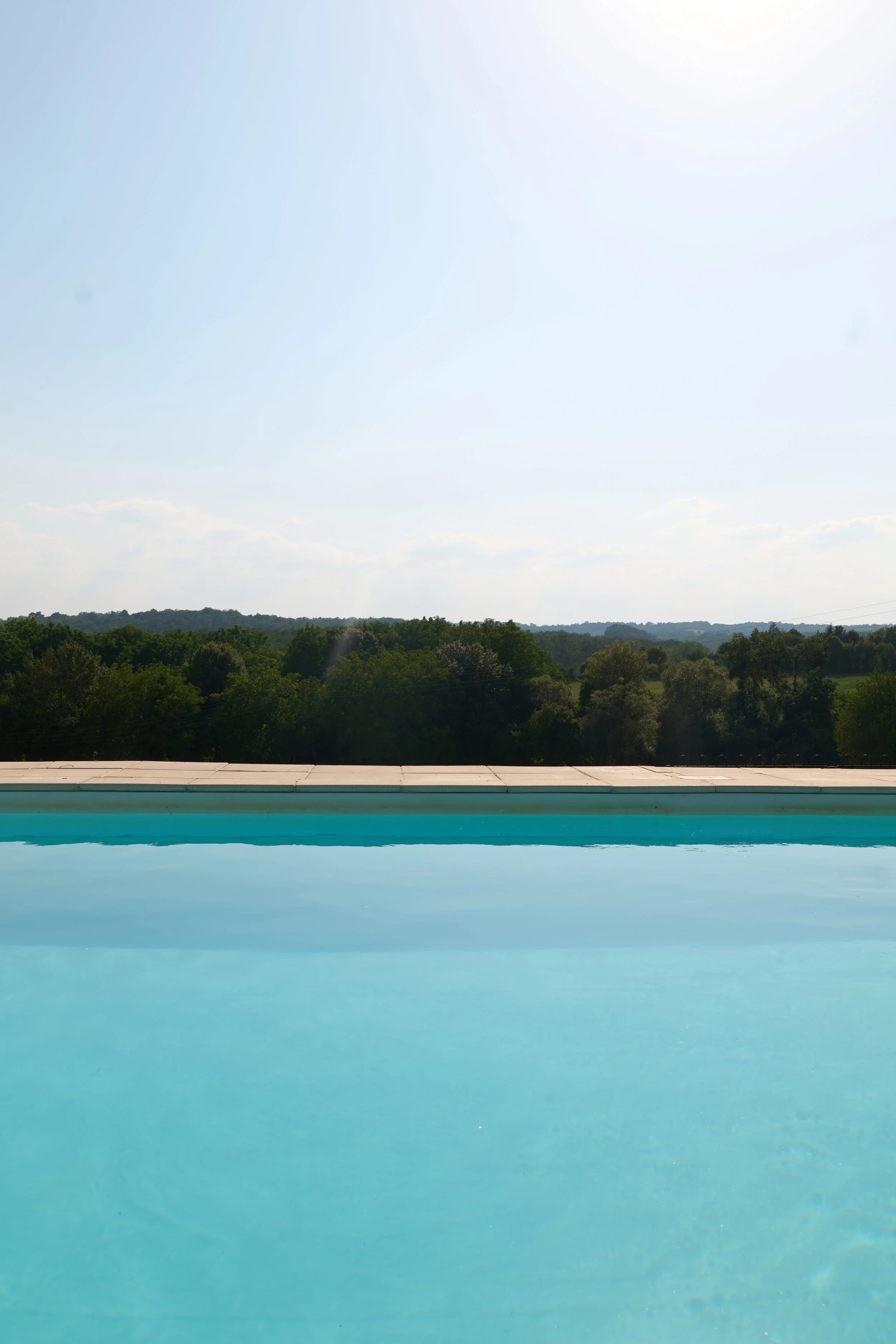 A swimming pool with a light blue surface, a white border, and a view of a distant tree-lined horizon under a partly cloudy sky.