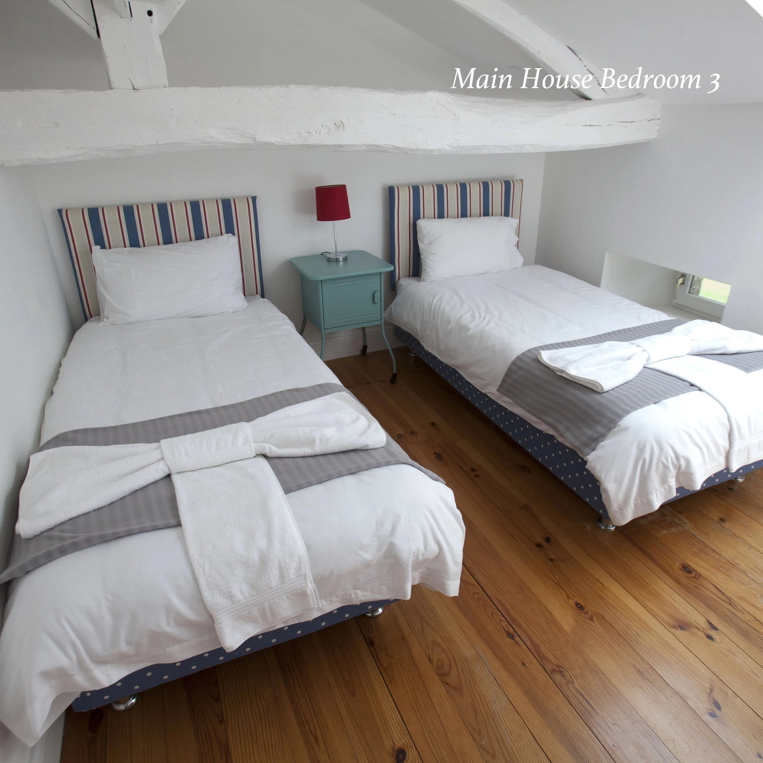 Two single beds with white bedding and striped headboards in a bedroom with white walls and a wooden floor. A red bedside lamp sits on a small teal nightstand between the beds, and a small window is visible on the right side. The room has an angled c