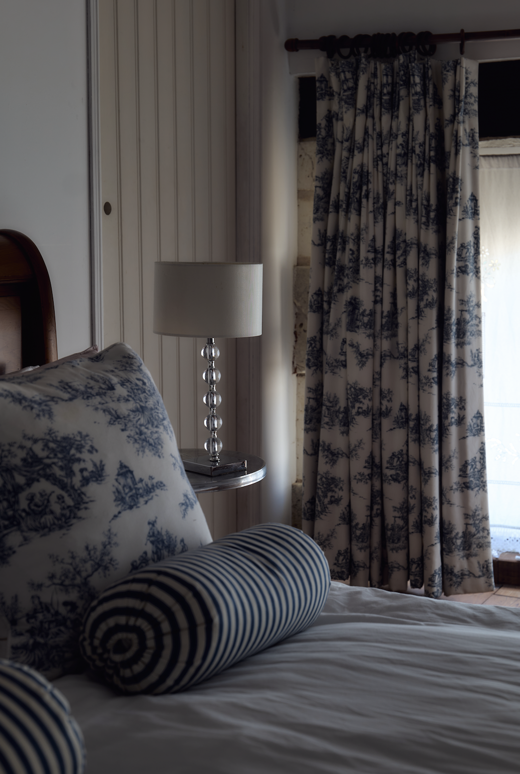 A cozy bedroom corner with a bed, blue and white toile pillow, striped bolster pillow, a bedside lamp with a glass bead base, and floral-patterned curtains near a window.
