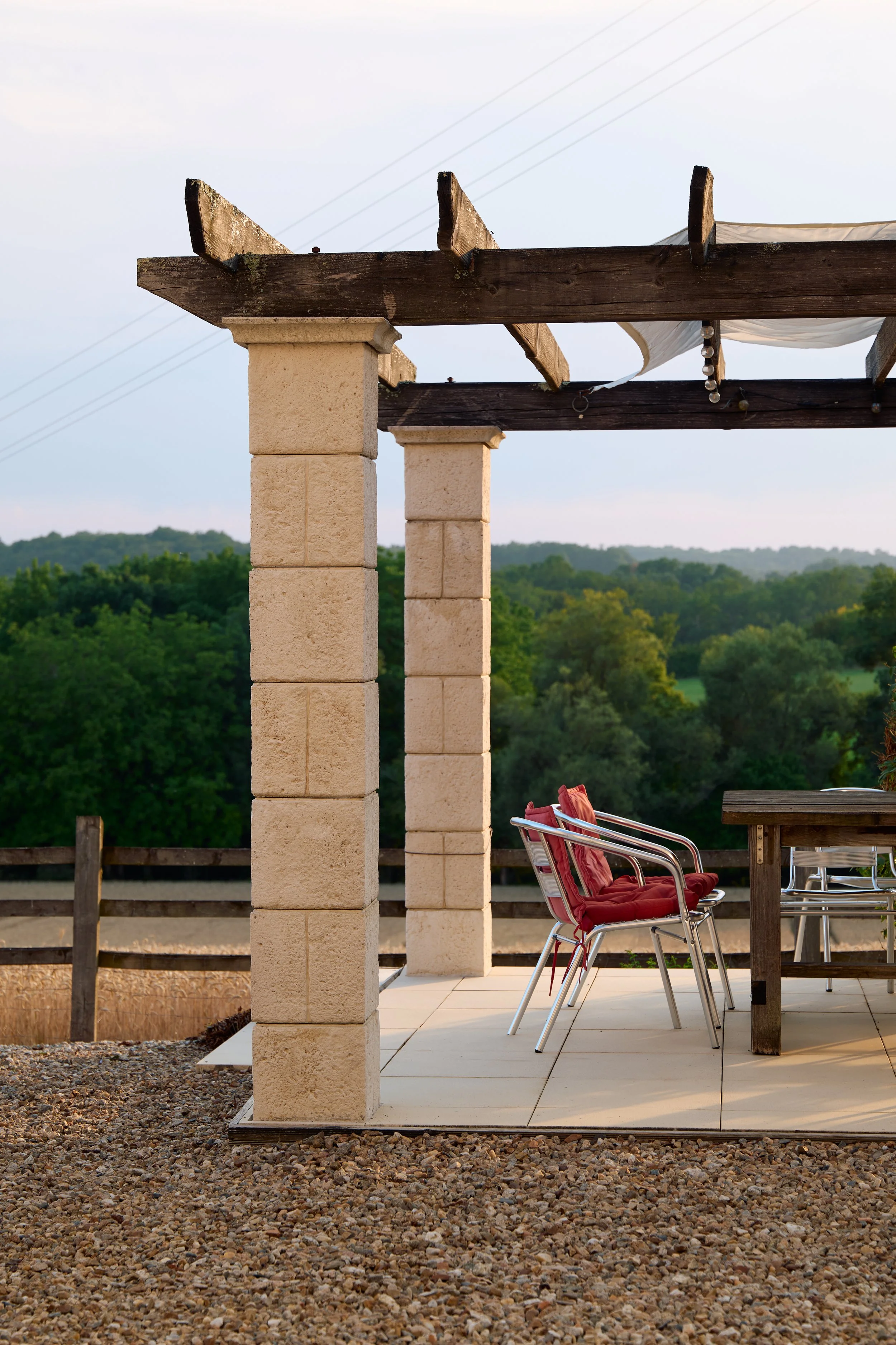 Outdoor patio with beige stone pillars, a wooden pergola structure, red chairs, and green trees in the background.