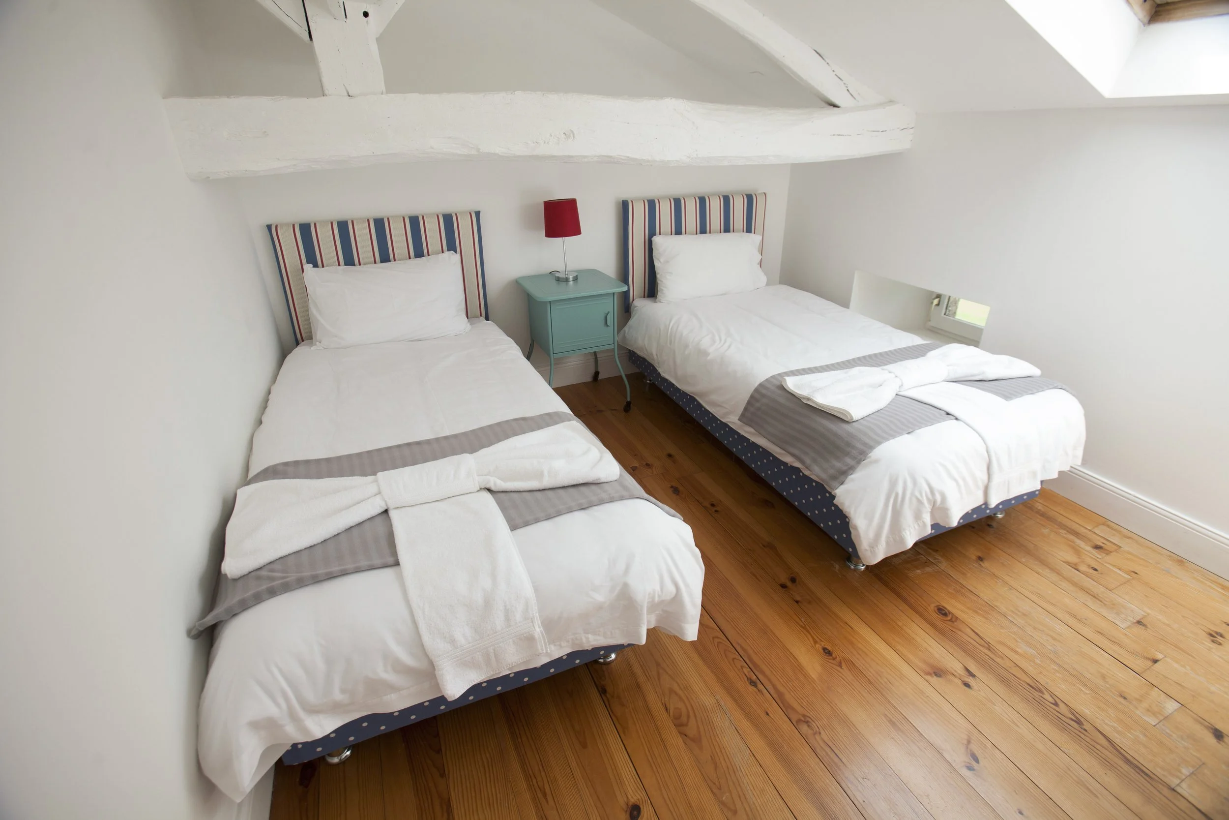 A bedroom with two single beds, each with white linens and gray runners, striped headboards in red, white, and blue, a teal nightstand with a red lamp, and a skylight window in a white-walled room with wooden flooring.