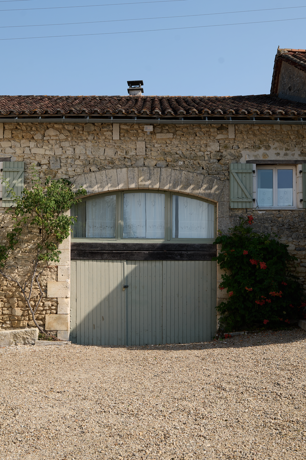 A stone house with a large arch-shaped window and a garage door below it, surrounded by greenery and red flowers, under a clear blue sky.