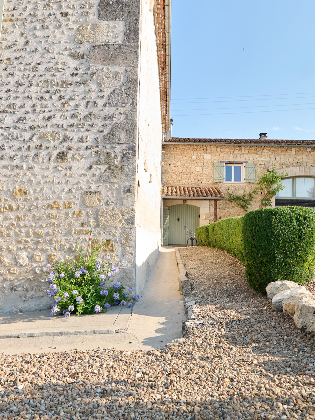 A stone building with a narrow concrete walkway, purple flowers, green bushes, and a small wooden door with a chair nearby, under a clear blue sky.