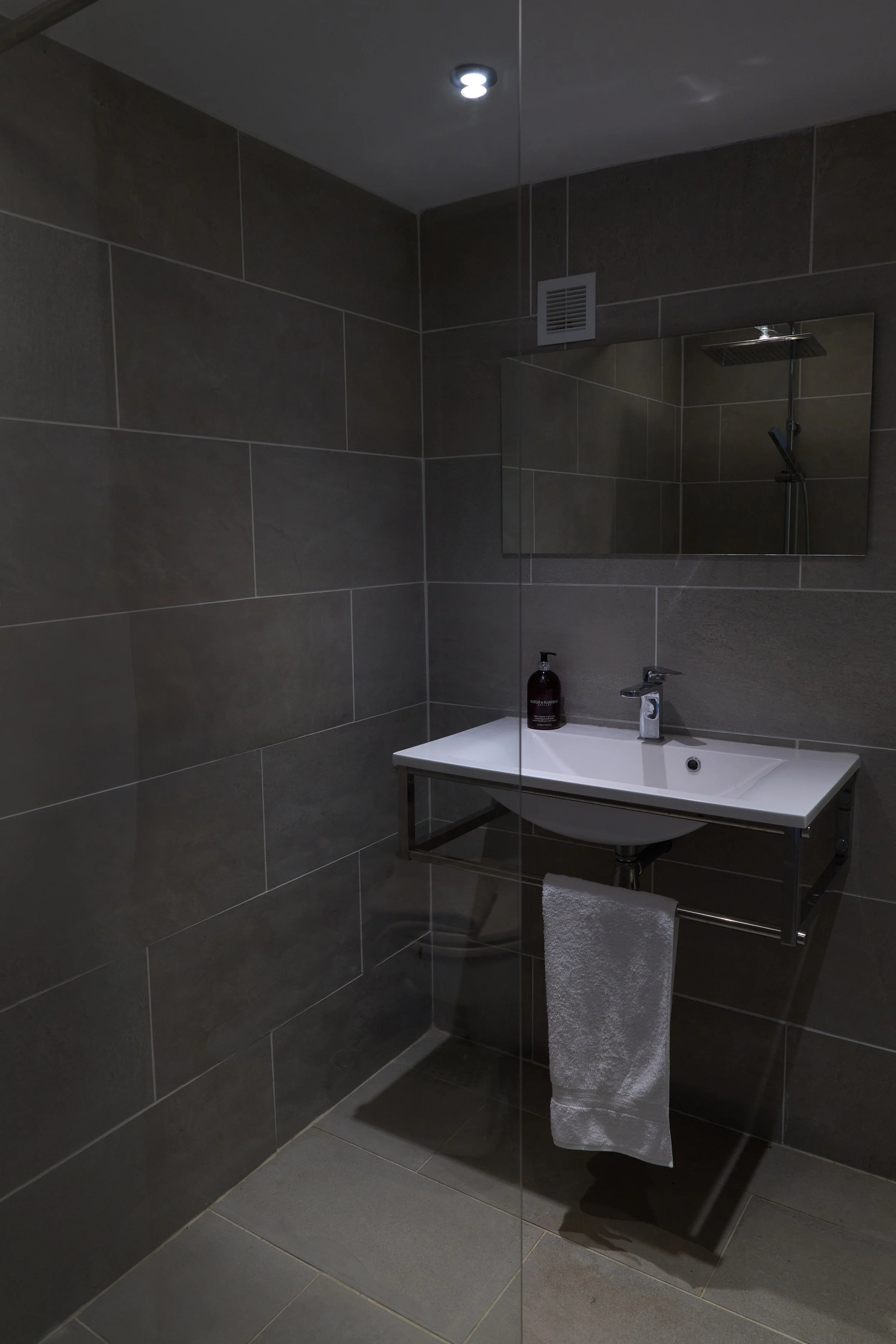Modern bathroom with dark gray tiled walls and floor, a white sink with a chrome faucet, a gray hand towel hanging from a rail, a bottle of soap on the sink, a wall-mounted mirror, a partially visible showerhead, a ceiling spotlight, and a ventilatio