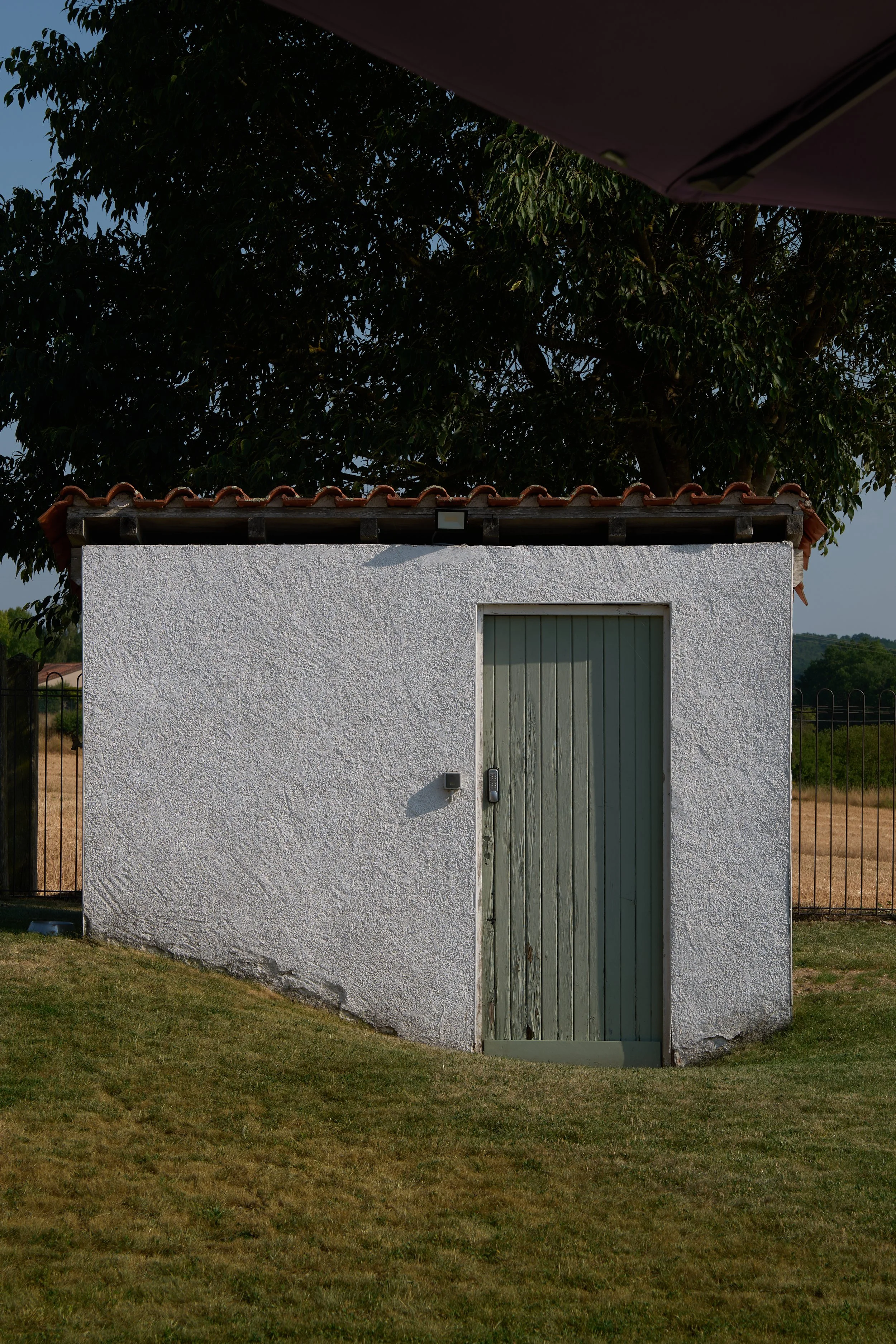 White stucco building with a tall, narrow, light green door and a small roof with red tiles. There is greenery and a tree behind the building, with a partly cloudy sky.
