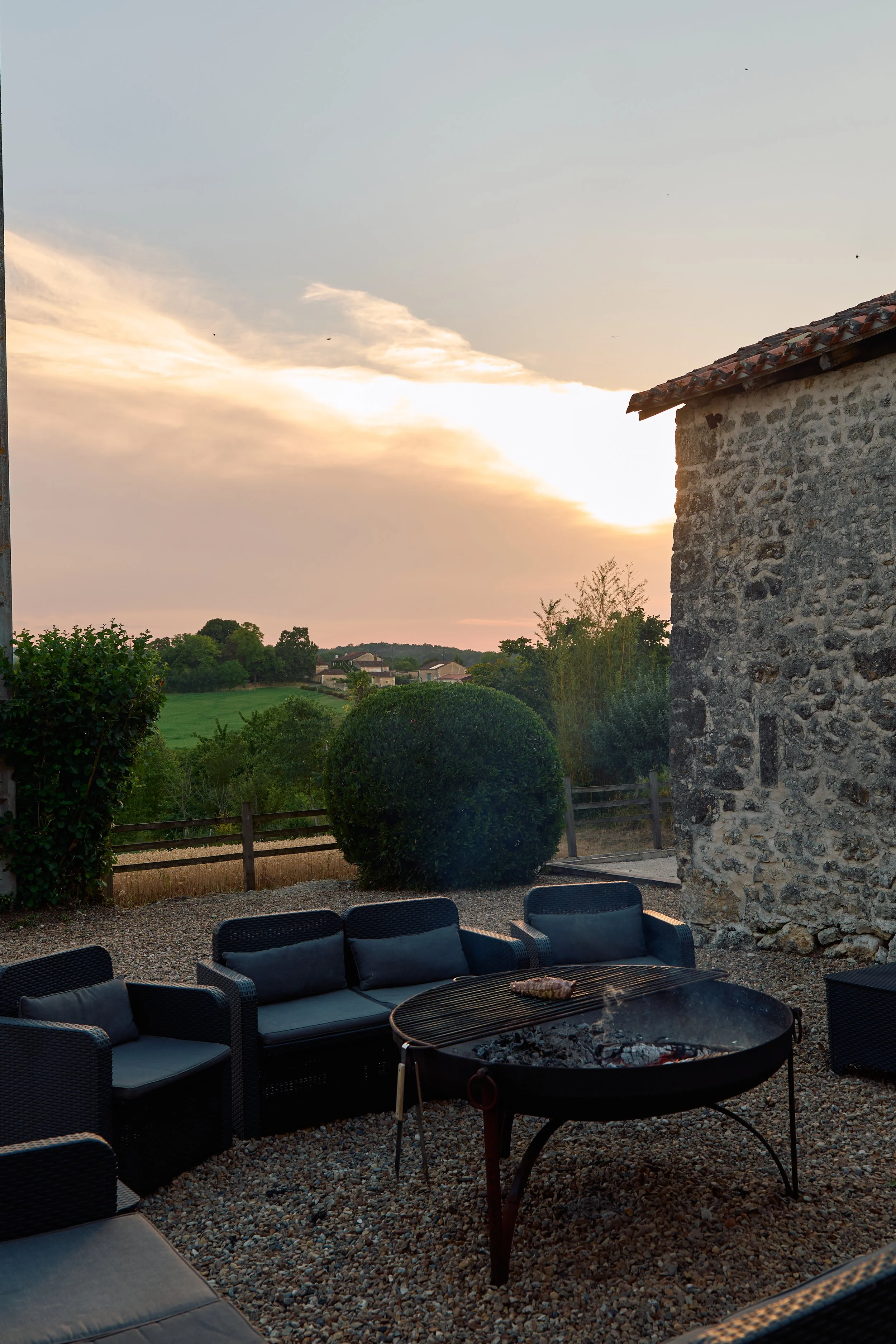 Outdoor seating area with black cushioned chairs around a fire pit, stone building on the right, green trees and houses in the distance, sunset sky with clouds.