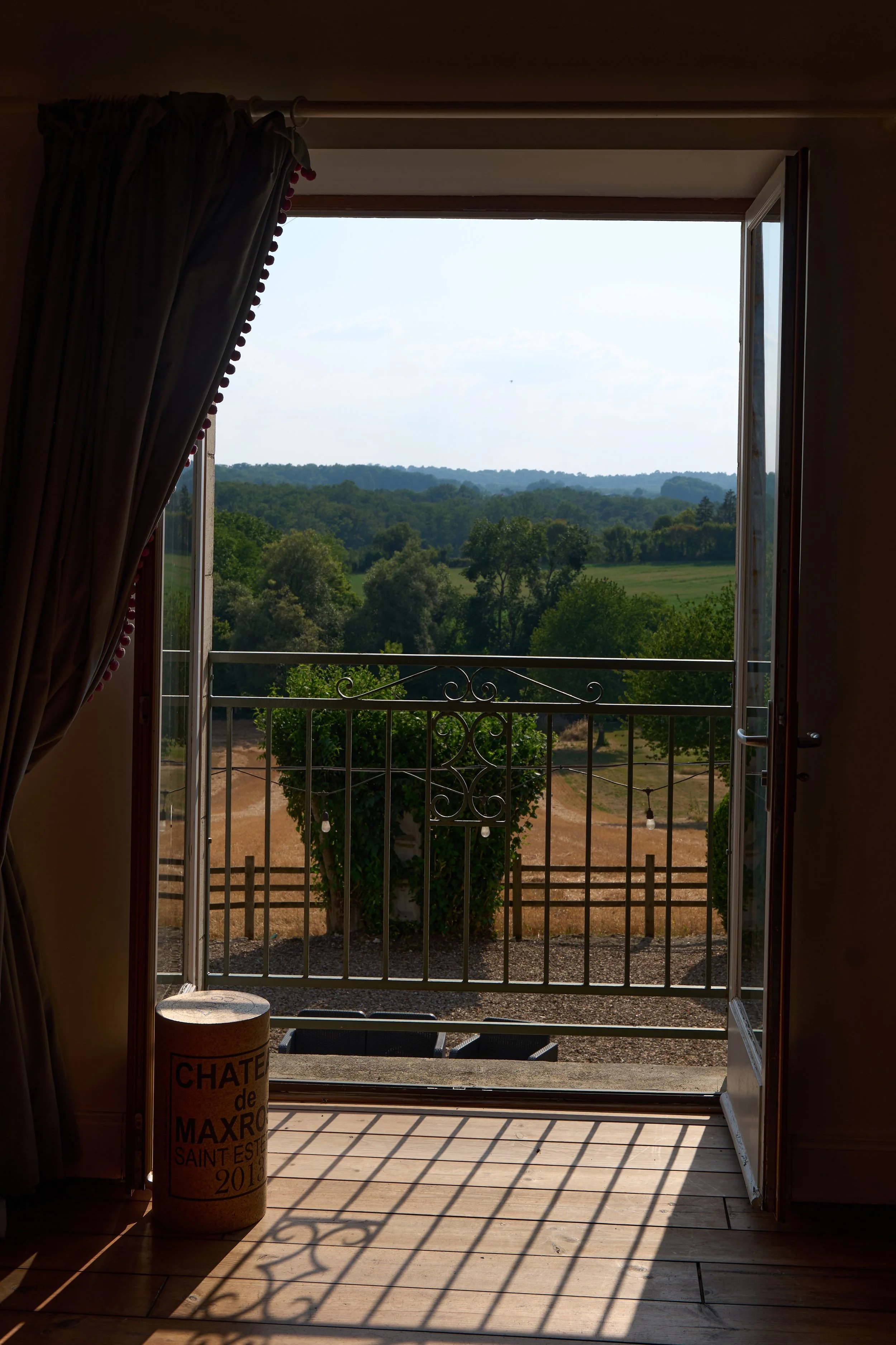 Open glass door leading to a balcony overlooking a green landscape with trees and rolling hills under a partly cloudy sky."