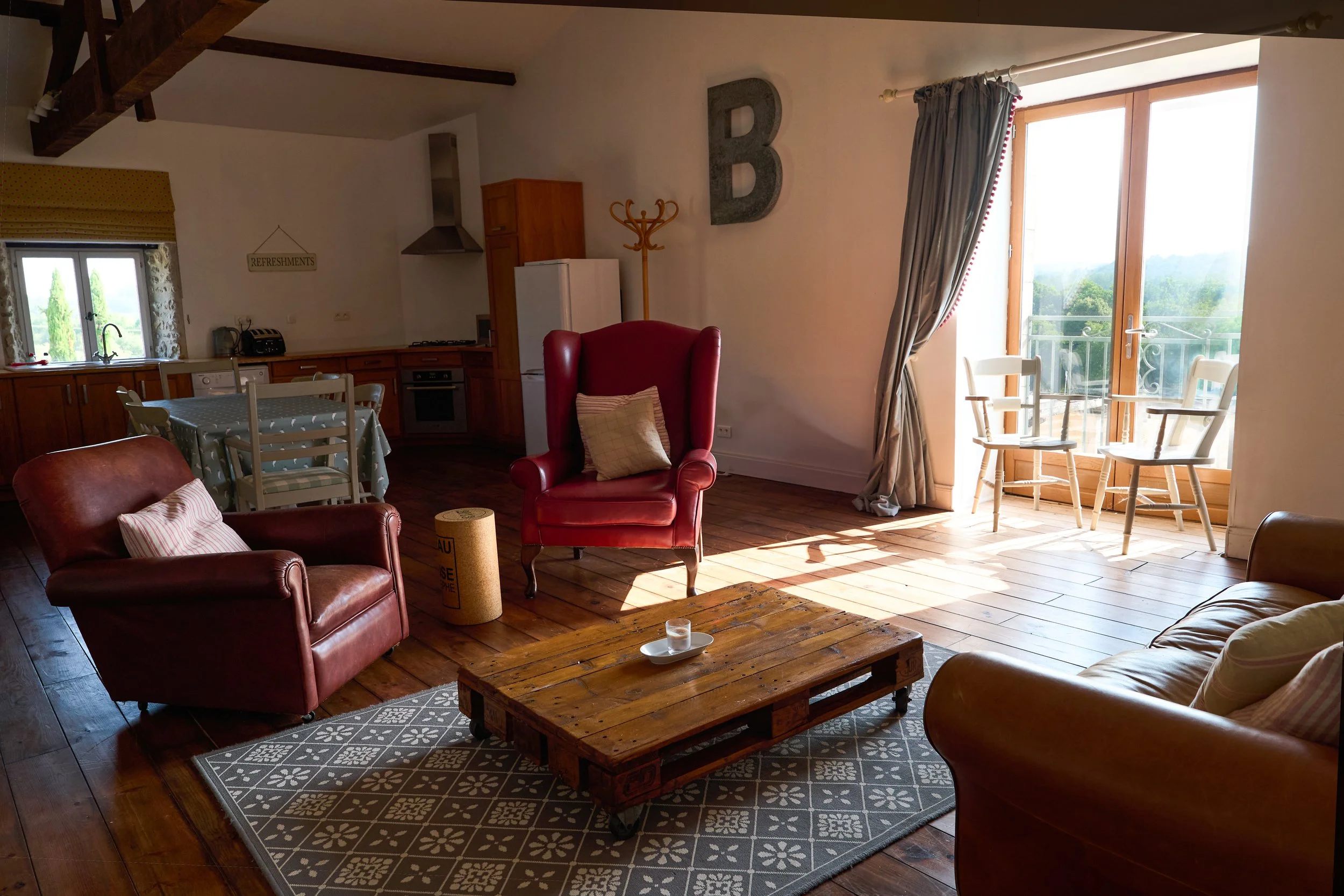 A cozy living room with wooden floors, a wooden coffee table on a patterned rug, and leather armchairs. There is a large window with curtains allowing sunlight, and a small balcony with two chairs outside. The kitchen area has wooden cabinets and a dining table with chairs, and decorative elements like a coat rack, a large letter 'B' on the wall, and a 'Refreshments' sign hanging above the window.