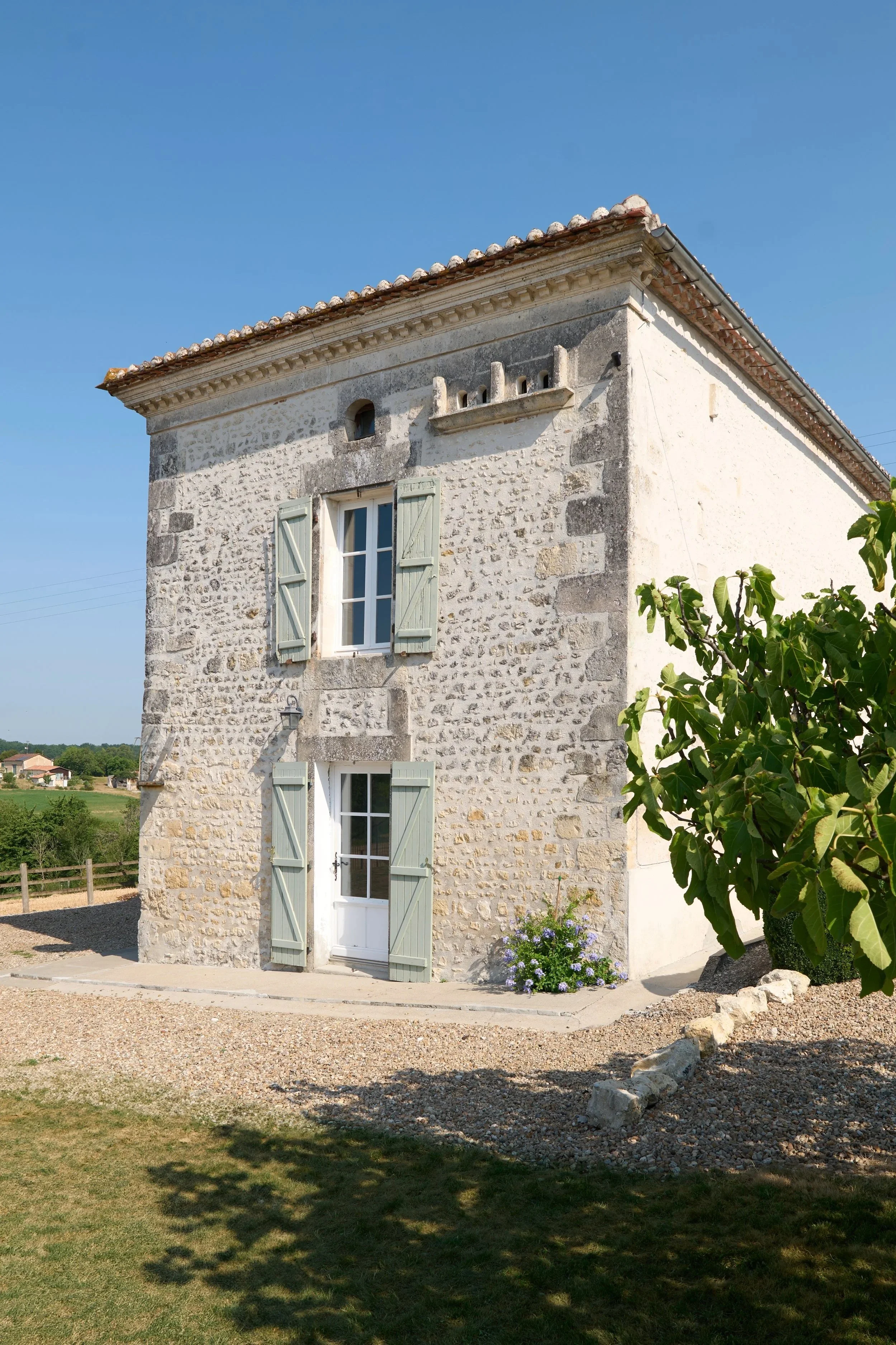 A stone house with pale blue shutters and a white door, surrounded by a garden with flowers and a tree, under a clear blue sky.