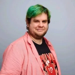 A man with green hair and a beard, wearing a black T-shirt with a red and white graphic, and a pink shirt over it, standing against a light gray background.