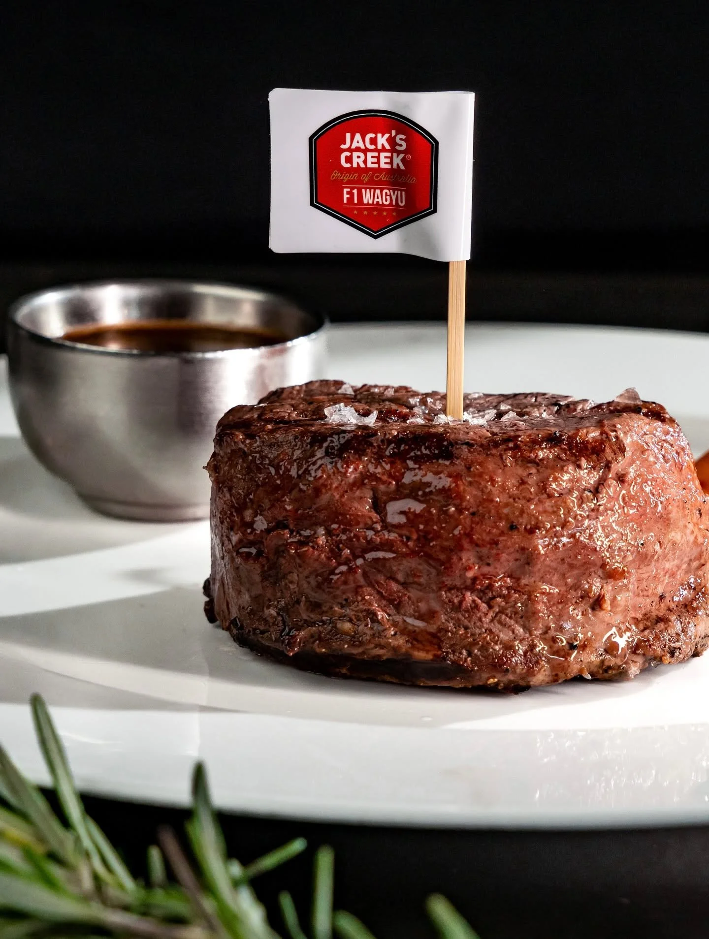 Cooked beef steak with a small flag reading "Jack's Creek F1 Wagyu" on top, placed on a white plate. A small bowl of sauce is in the background, and some herbs are visible in the foreground.