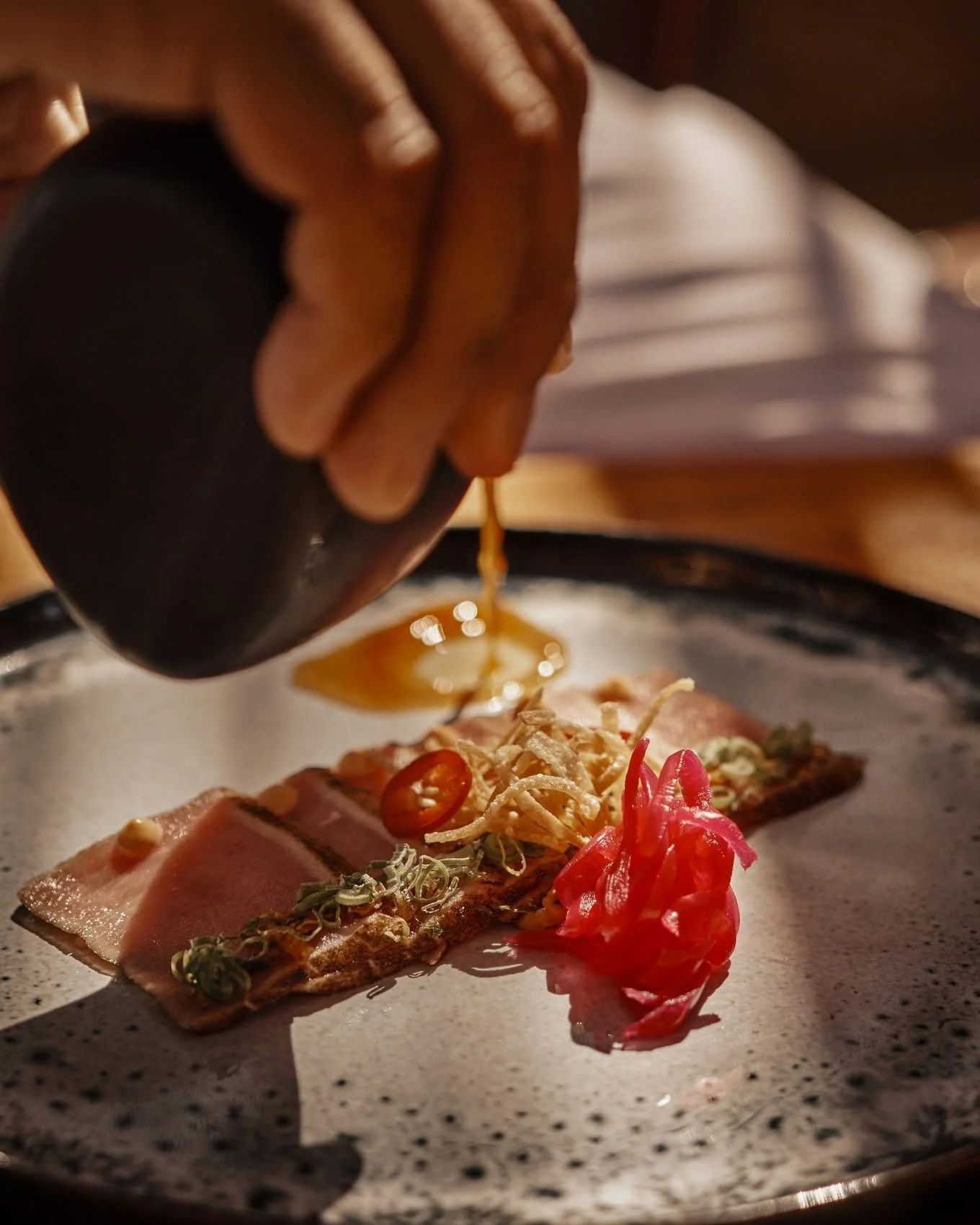 A person's hand pouring sauce over a dish with slices of beef, pickled ginger, and garnishes on a plate.