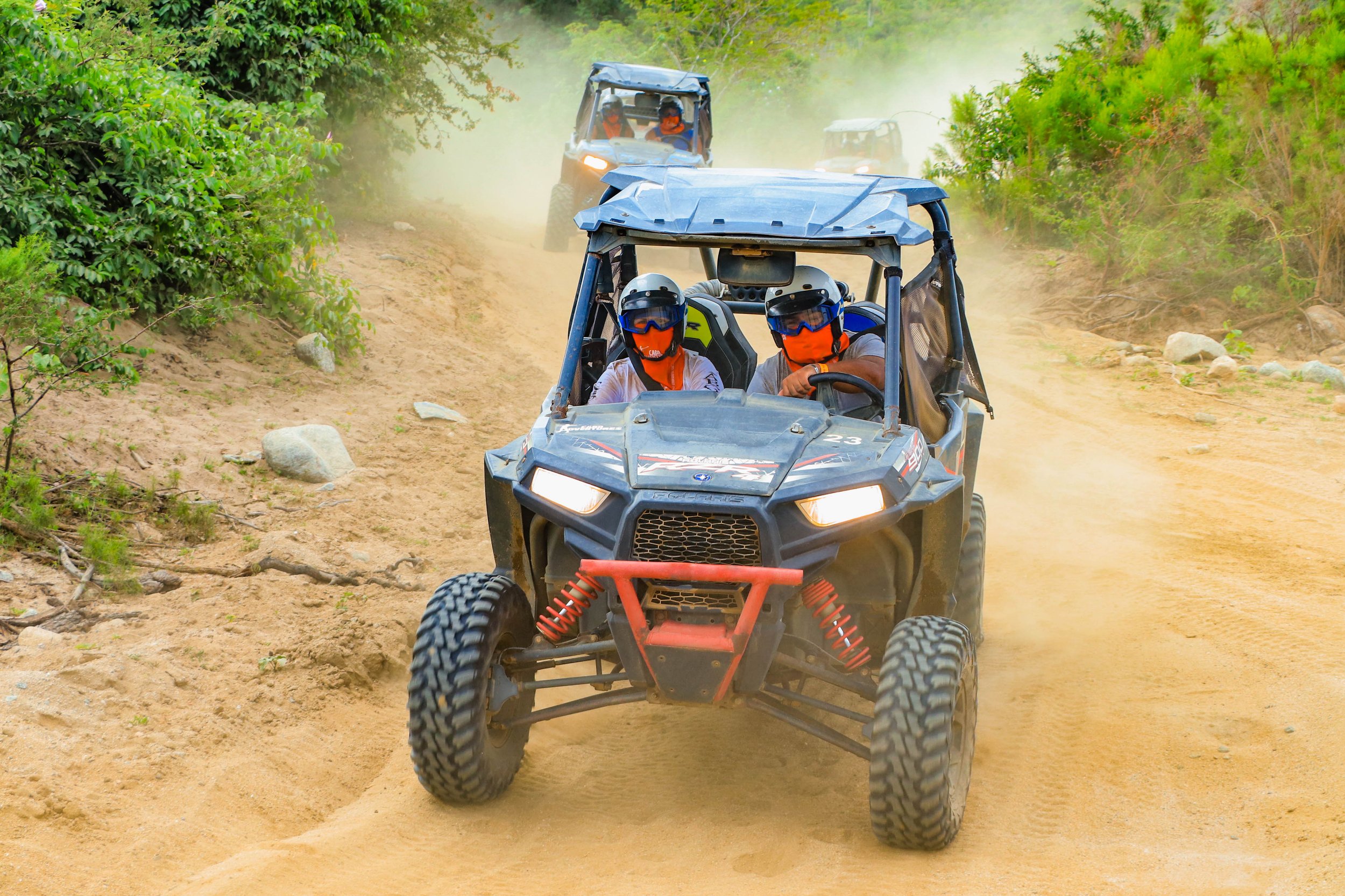Two all-terrain vehicles (ATVs) driving on a dirt trail through a green wooded area, with dust clouds behind them. The drivers are wearing helmets and protective gear.