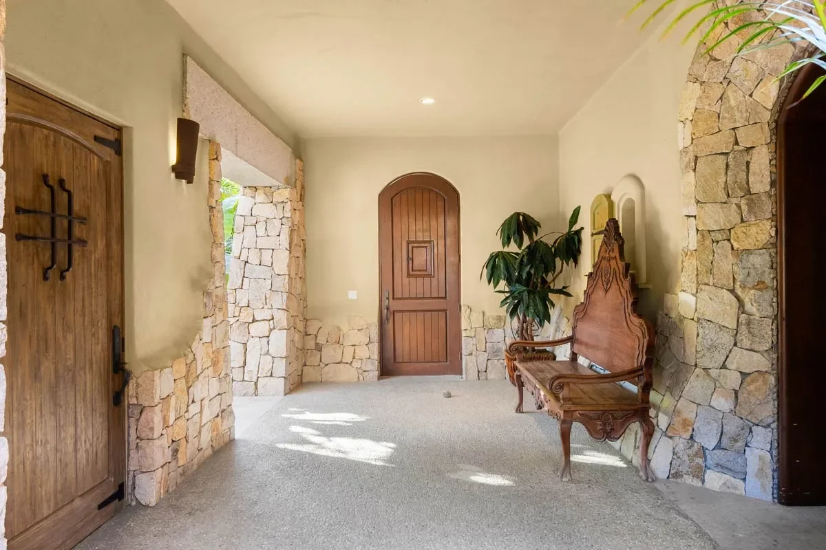 An entrance hallway with stone walls, a wooden door, a wooden bench, a potted plant, and natural light coming through an archway on the left.