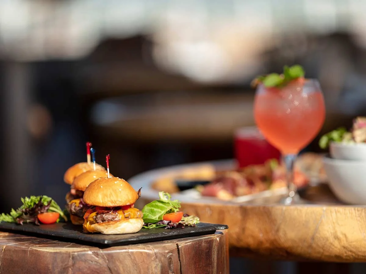 Three mini burgers with cheese, tomato, and lettuce on a black serving slate, garnished with fall salad greens, placed on a rustic wooden table.