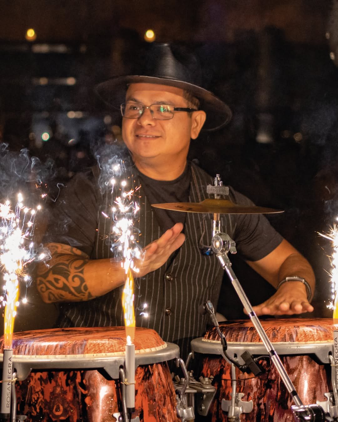 A man in a black hat and glasses playing drums surrounded by sparklers at night.
