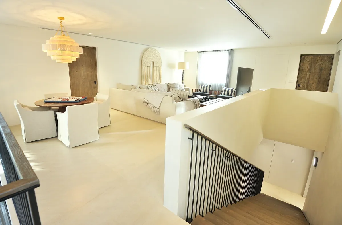 Bright and modern living room with white walls, a large sofa, a TV, a dining table with chairs, and a staircase leading downstairs.