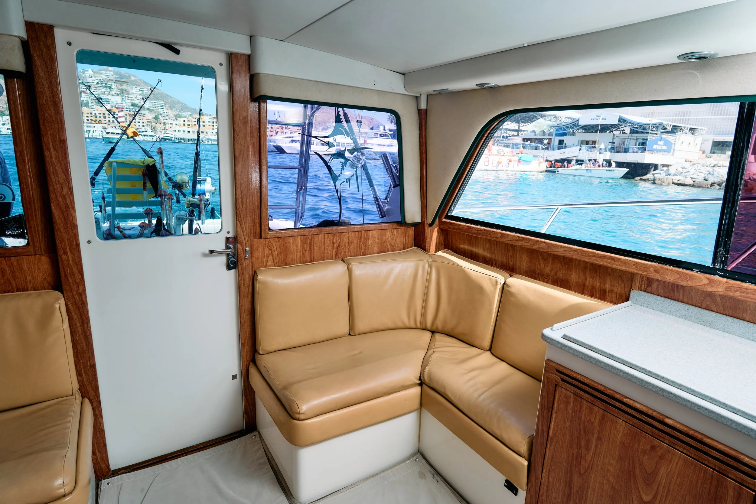 View of a boat's interior with beige leather seating, wooden trim, and windows showing a marina with boats and buildings in the background.