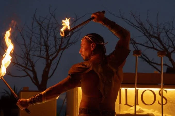 A performer holding a flaming torch in each hand while dancing or performing outdoors at night with leafless trees in the background.