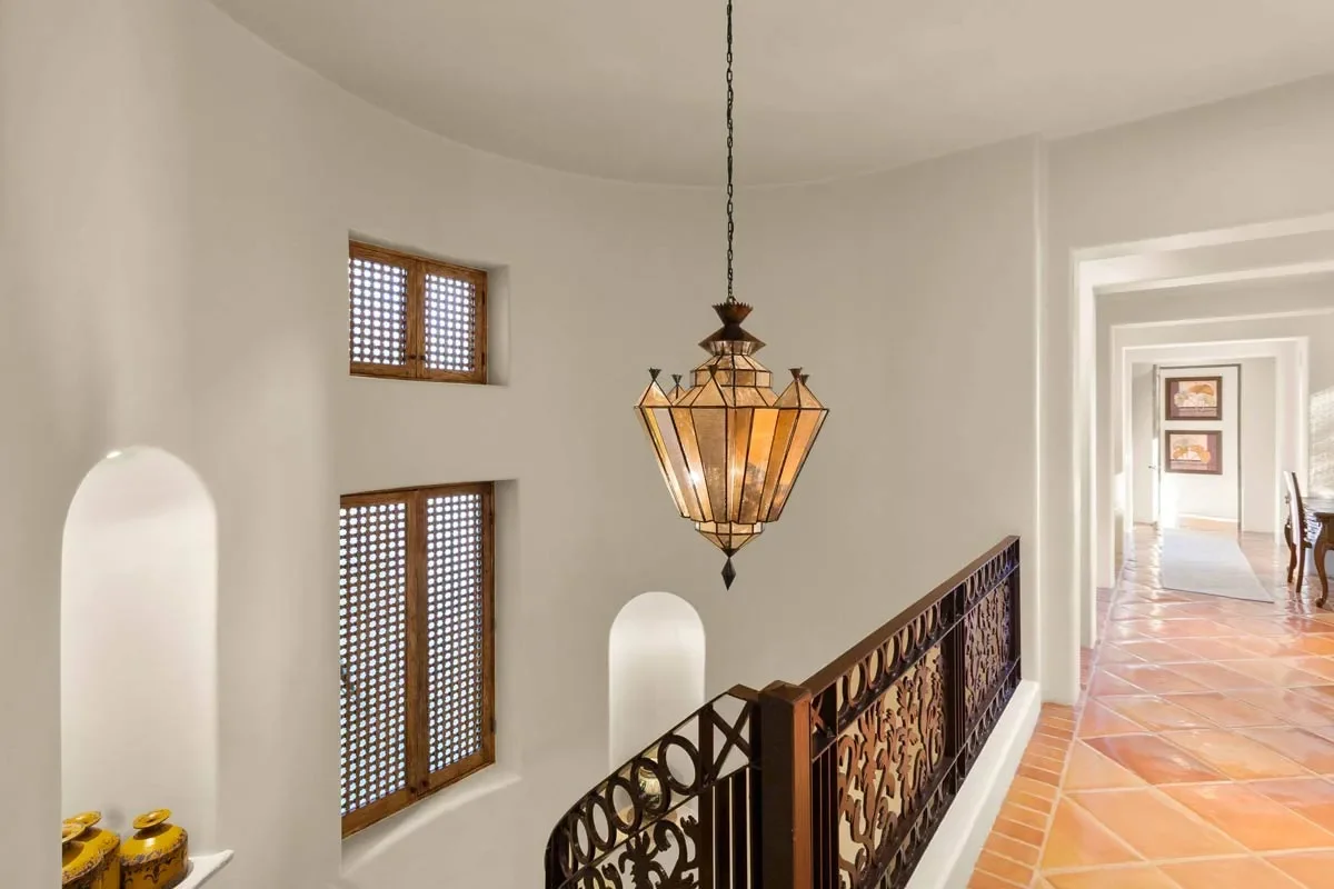 Interior view of a hallway with white walls, wooden windows with lattice design, terracotta tile flooring, an ornate hanging lantern, and a decorative wrought iron railing.
