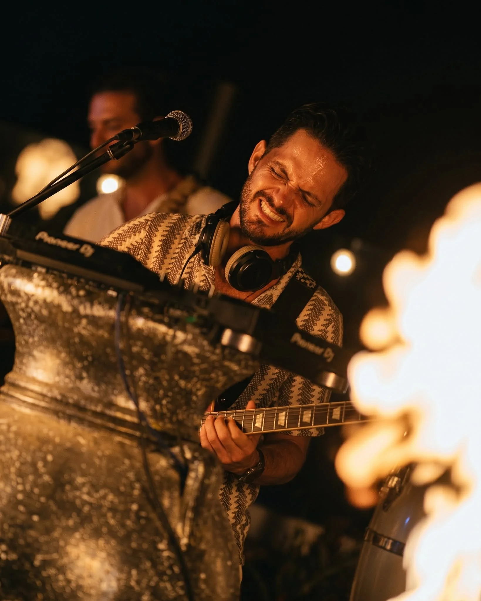 Musician playing guitar and singing with a passionate expression, captured in warm, dim lighting, with a microphone near his mouth and a person in the background.
