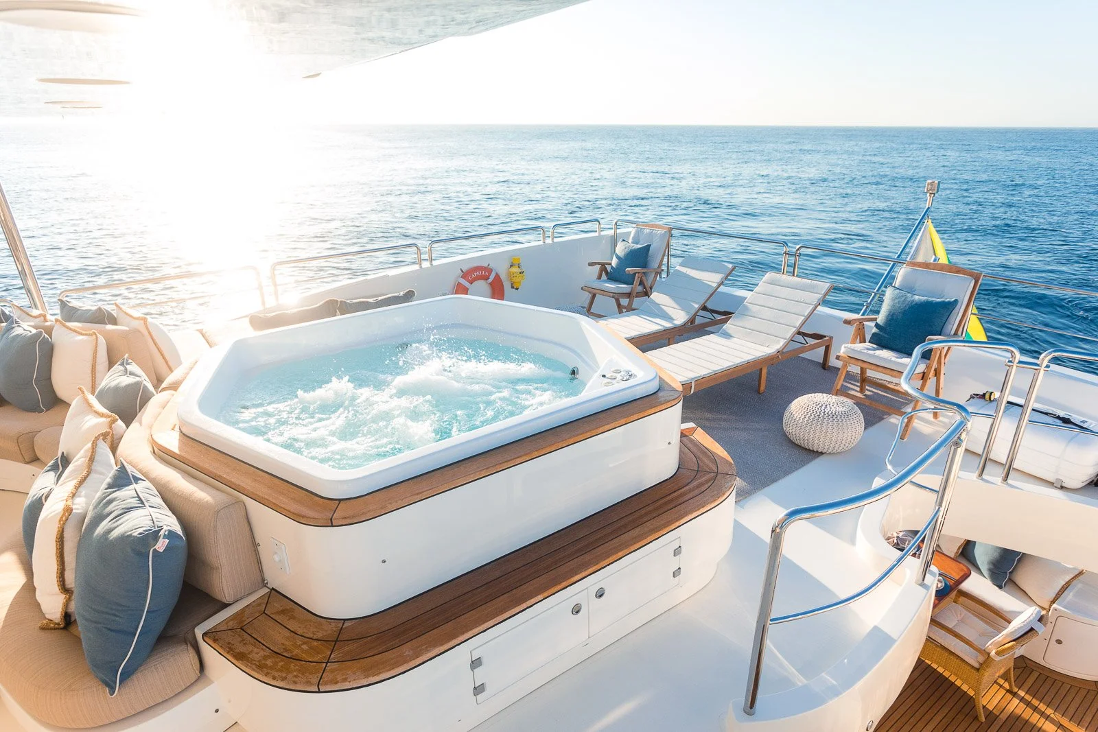 Luxury yacht deck with hot tub, lounge chairs, pillows, on-deck seating, and ocean view in the background.