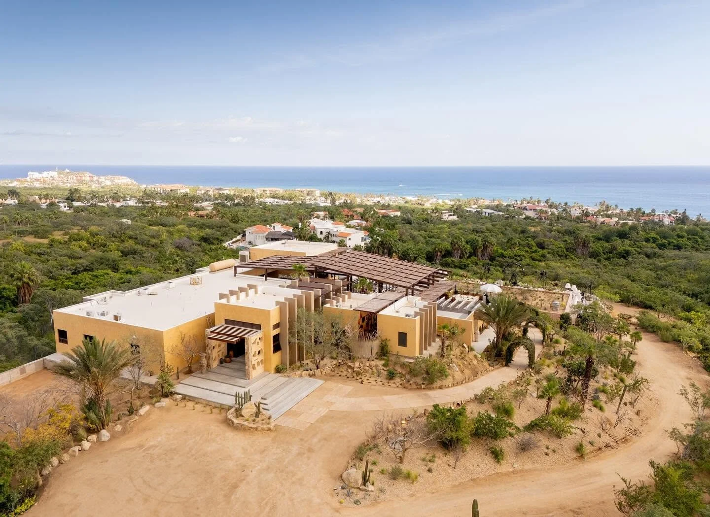 An aerial view of a modern desert-style house with a yellow exterior, surrounded by desert landscaping including palm trees and cacti, with a dirt driveway leading up to it, overlooking a lush green area and a distant ocean under a clear blue sky.