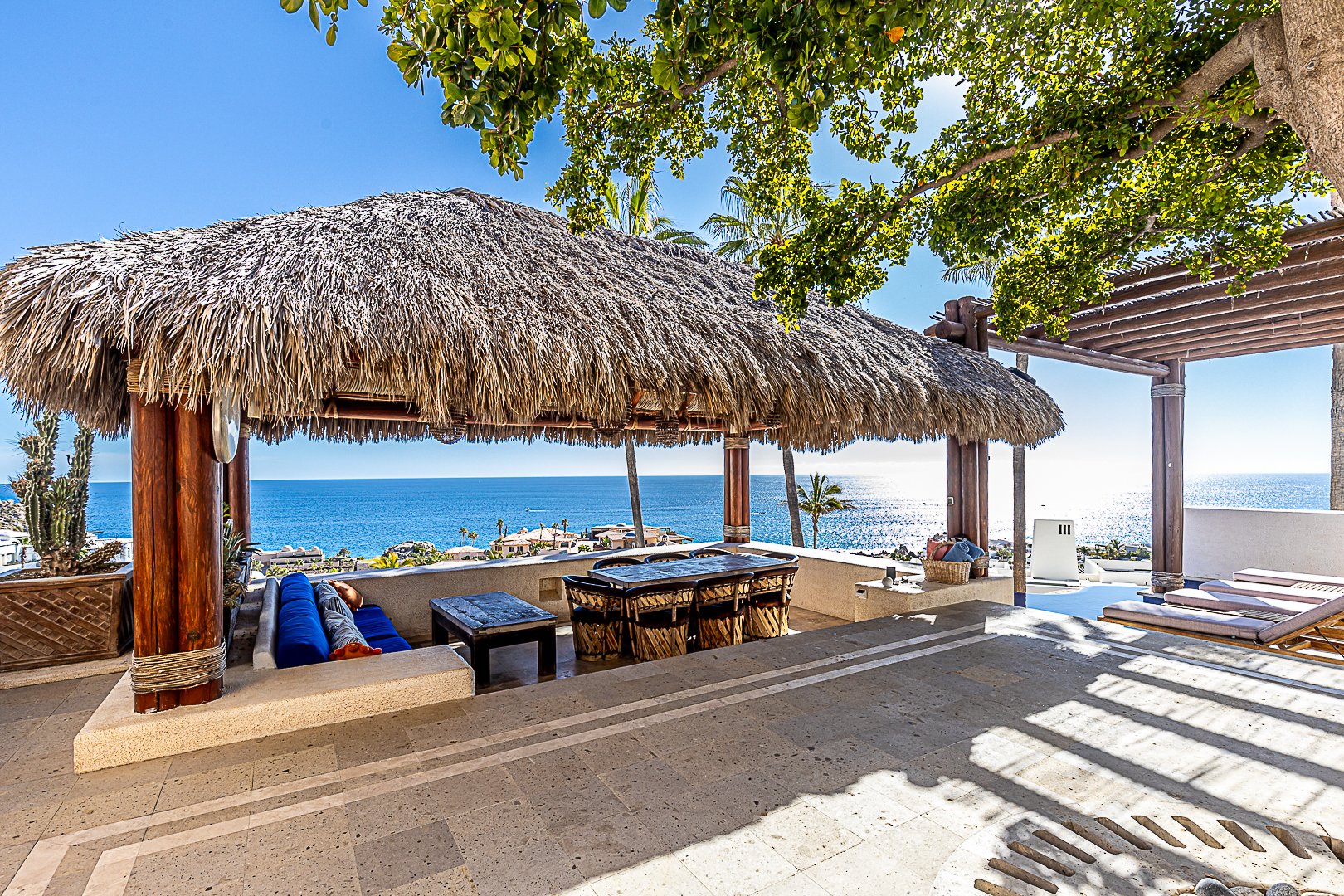 A tropical patio overlooking the ocean, featuring a thatched-roof palapa with seating and a view of the water, palm trees, and a clear blue sky.