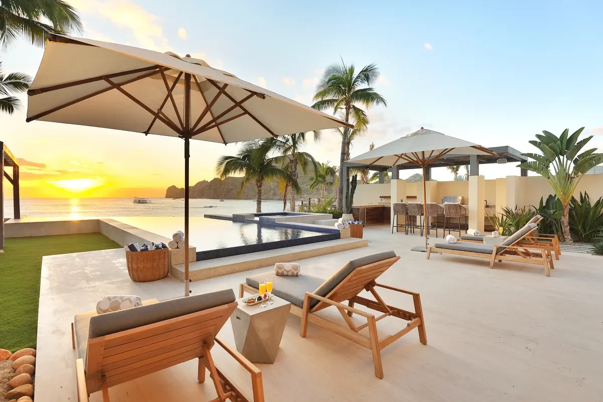 Luxury outdoor pool area at sunset with palm trees, lounge chairs, umbrellas, and ocean view in the background.