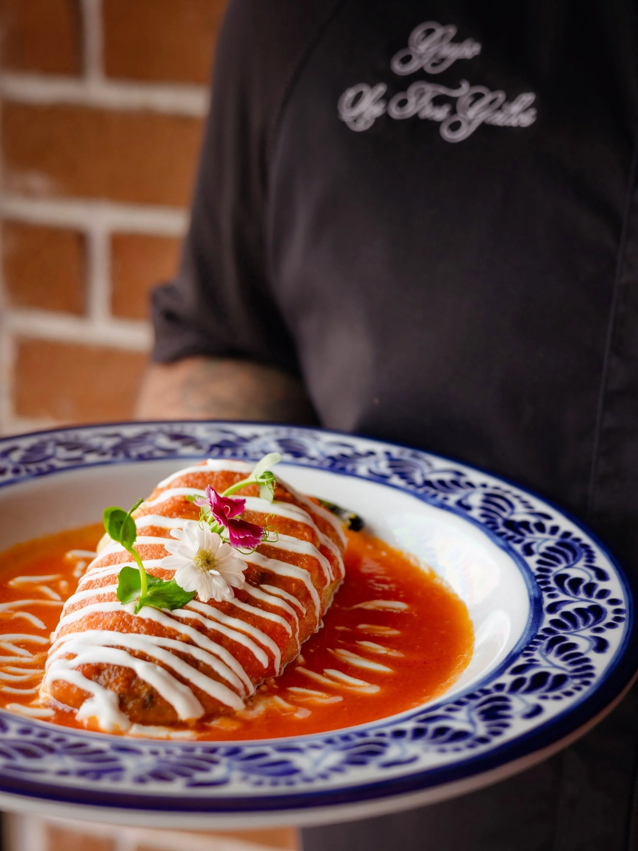 Close-up of a dish with a rolled item covered in white sauce, garnished with edible flowers, served on a blue and white patterned plate with a red sauce underneath.