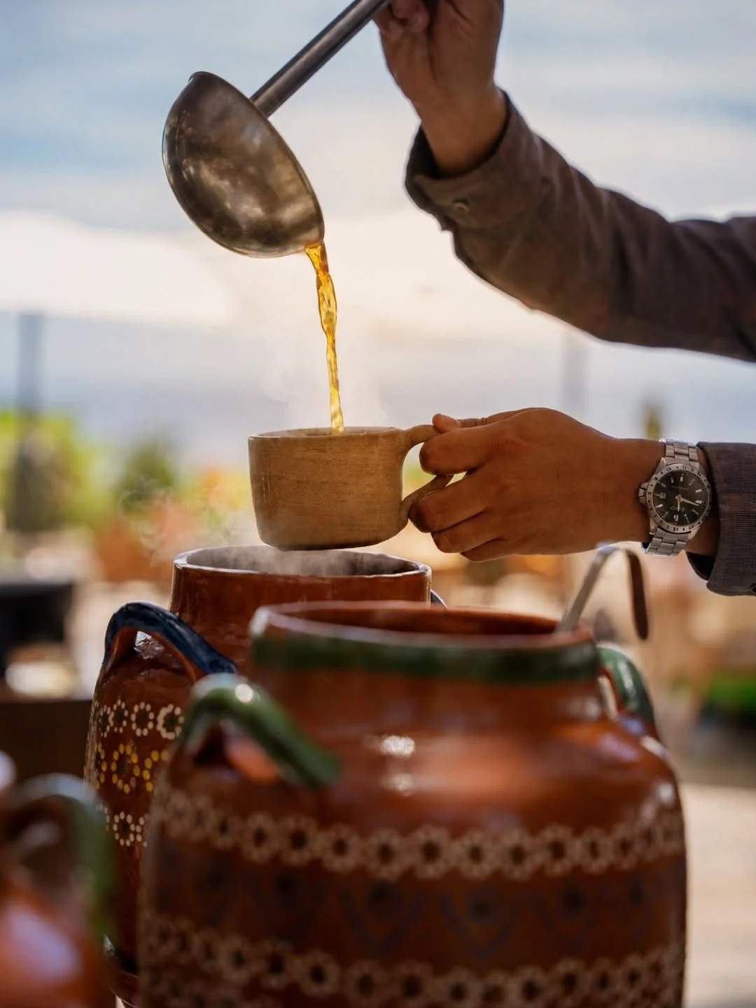 A person pours hot coffee from a metal pitcher into a ceramic cup outdoors, with traditional decorative pottery in the foreground and a cloudy sky in the background.