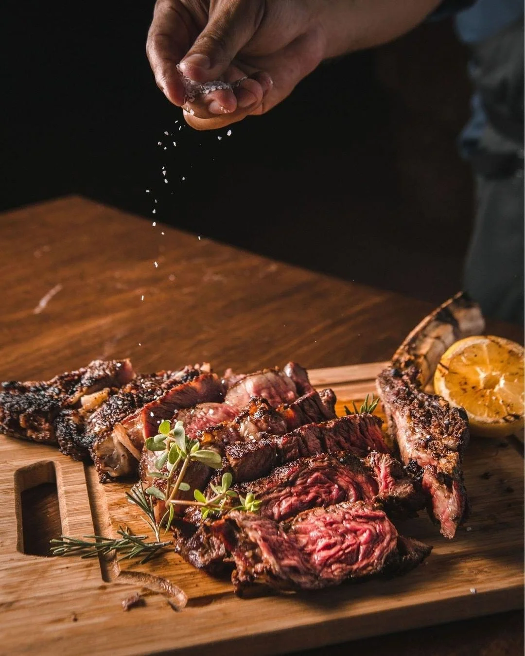 Person sprinkling salt on a variety of grilled steaks and a lemon half on a wooden serving board.