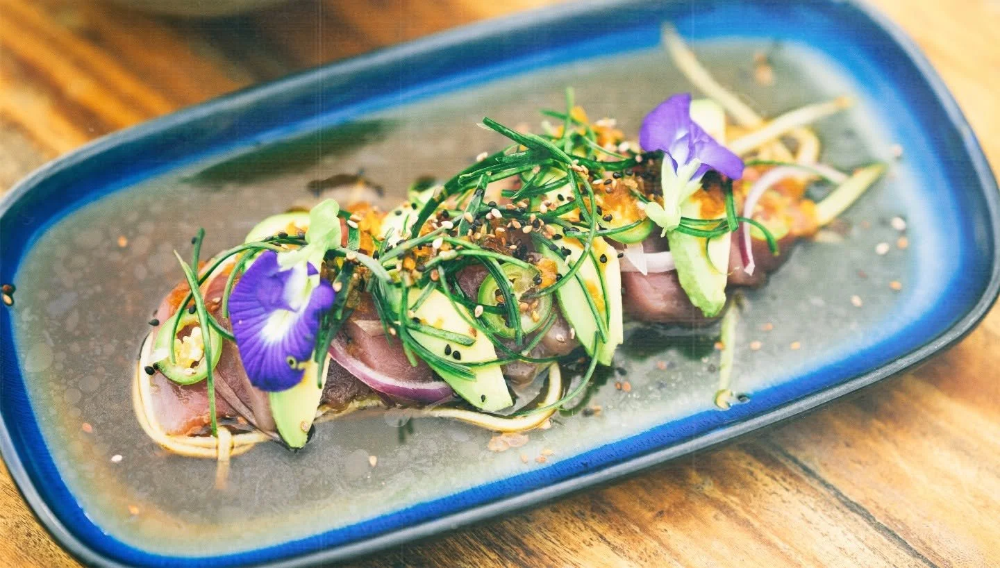 Sliced raw fish topped with cucumber slices, red onion, chives, edible flowers, sesame seeds, and spices on a blue oval plate.