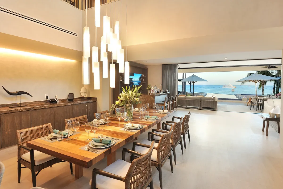 Modern dining room with a large wooden table, set with plates, glasses, and napkins, overlooking a beach with umbrellas and a boat in the ocean.