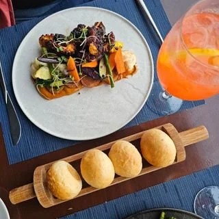 A white plate of mixed salad with greens, carrots, and other vegetables, a wooden serving board with five round fried appetizers, and a glass of orange cocktail with ice.