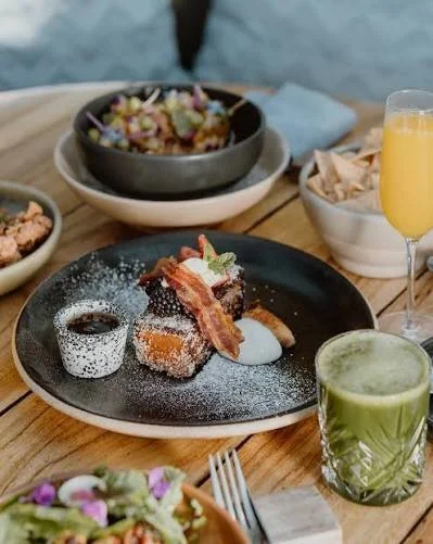 A black plate with dessert topped with whipped cream and bacon, accompanied by a small cup of sauce, on a wooden table. Surrounding are bowls with salad, chips, a green juice, and a glass of orange juice.