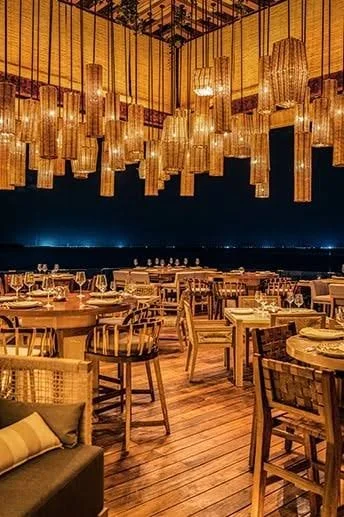Upscale restaurant with warm lighting and hanging bamboo lanterns, wooden tables and chairs, and a dark ocean or city view in the background.