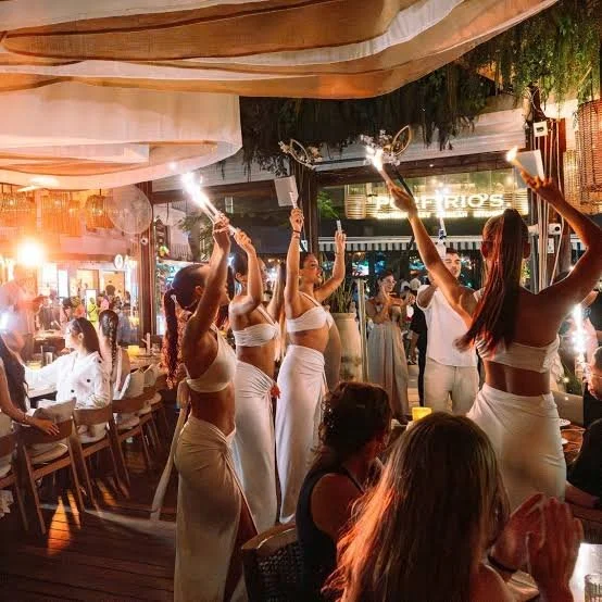 Group of women in white dresses dancing and holding sparklers at a lively indoor event with seated guests and warm lighting.