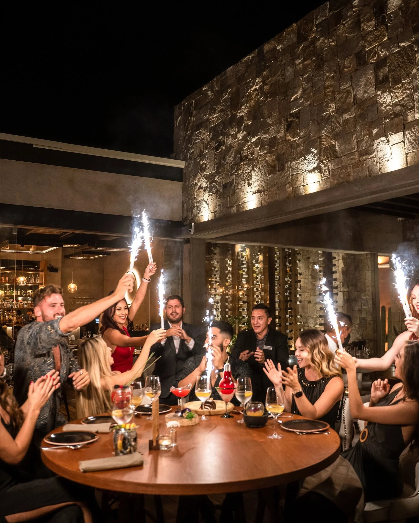 A group of people celebrating at a dinner party with sparklers, drinks, and food in a cozy restaurant with a stone wall and wine racks in the background.