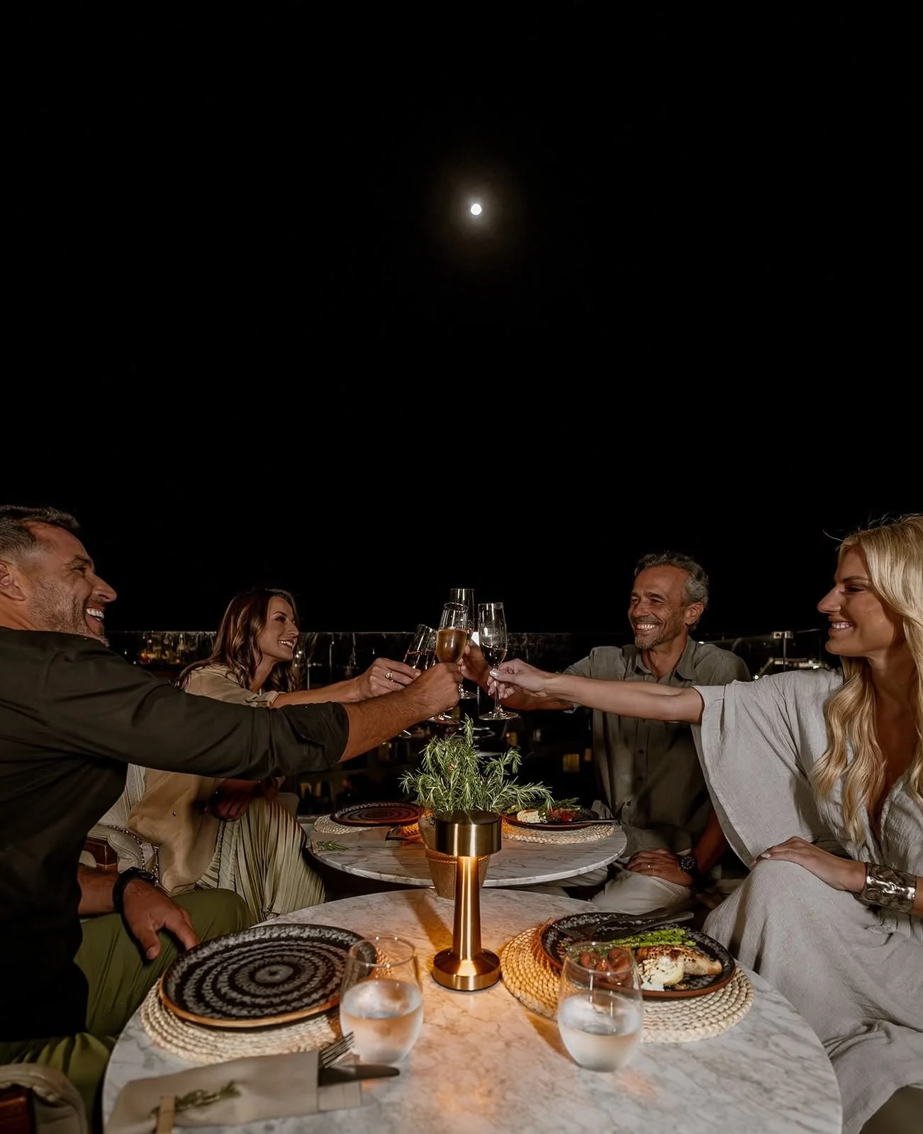 A group of five people enjoying a dinner party outdoors at night under a full moon, raising their glasses in a toast and smiling.
