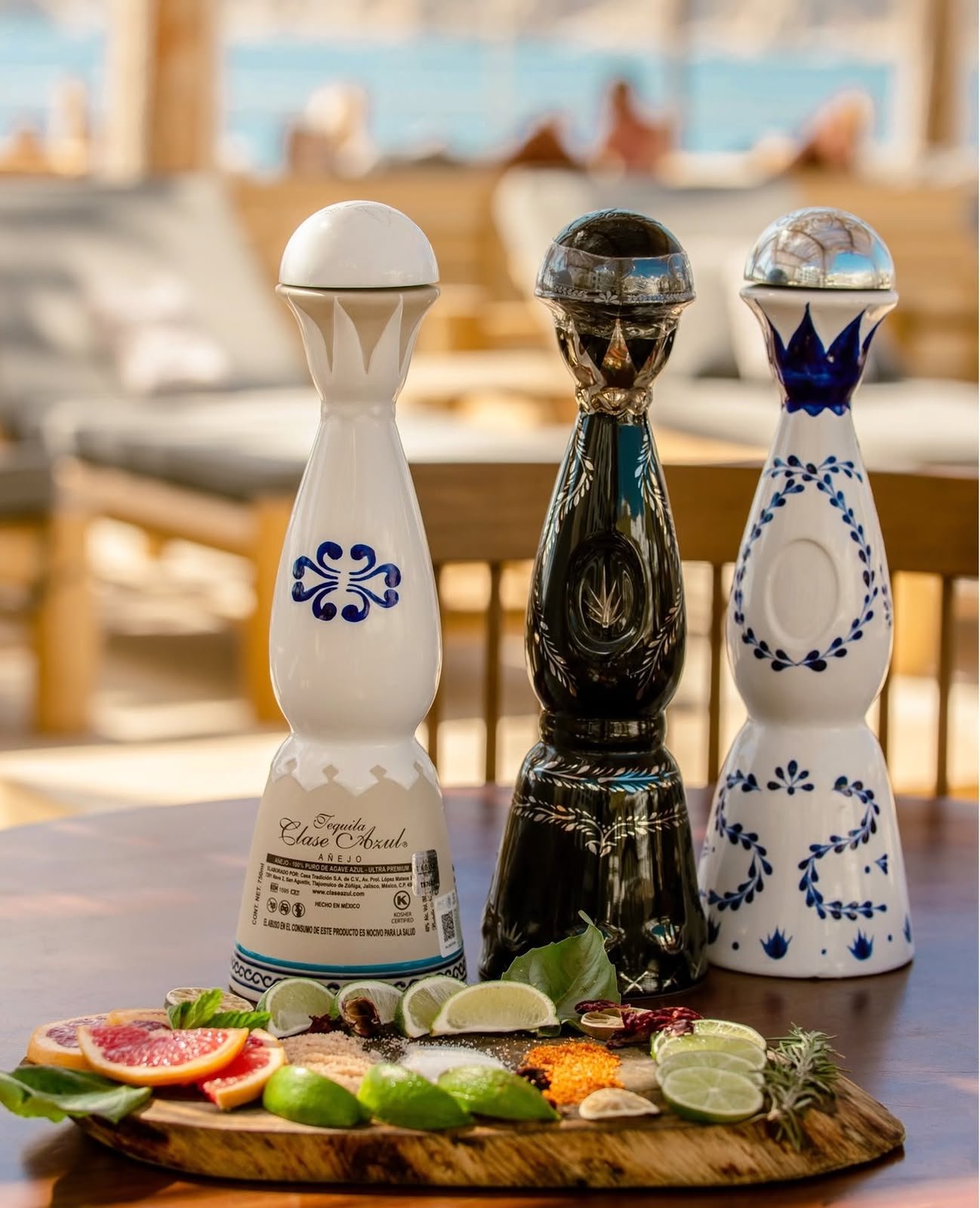Three decorative bottles of tequila on a wooden table with lime wedges, grapefruit slices, and herbs in front, set in a bright, indoor space with a wooden chair in the background.