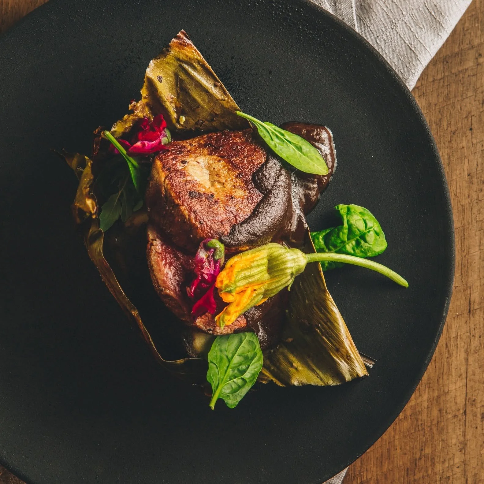 A cooked piece of meat served on a black plate, garnished with fresh greens, a yellow zucchini, and a roasted vegetable, with a white napkin and wooden surface nearby.