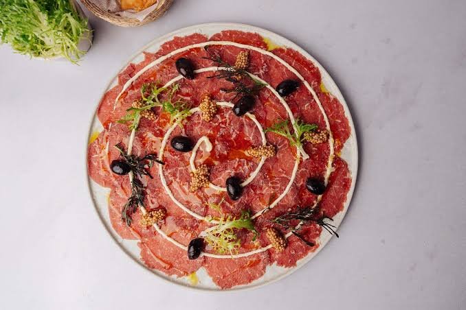 A round plate of assorted raw meat slices, garnished with black olives, mustard seeds, green herbs, and decorative white sauce swirls on top.