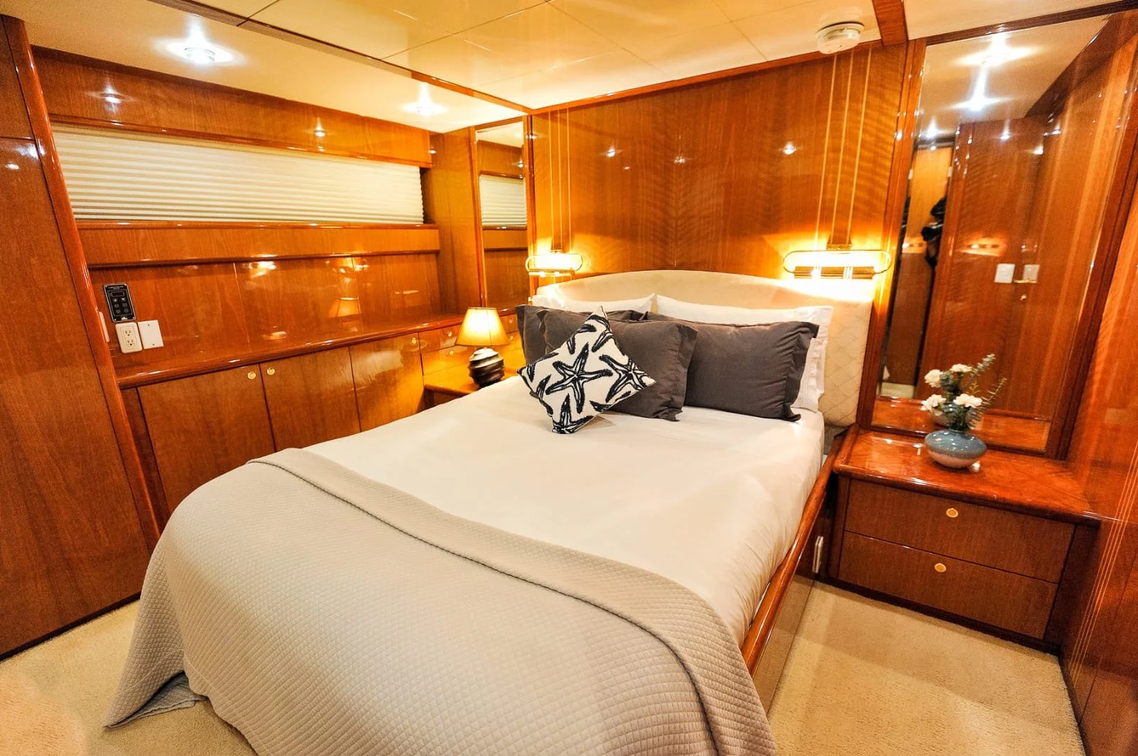 A cozy bedroom with wood-paneled walls, a made bed with white bedding and decorative pillows, a bedside lamp, and a small vase of flowers on a wooden nightstand.