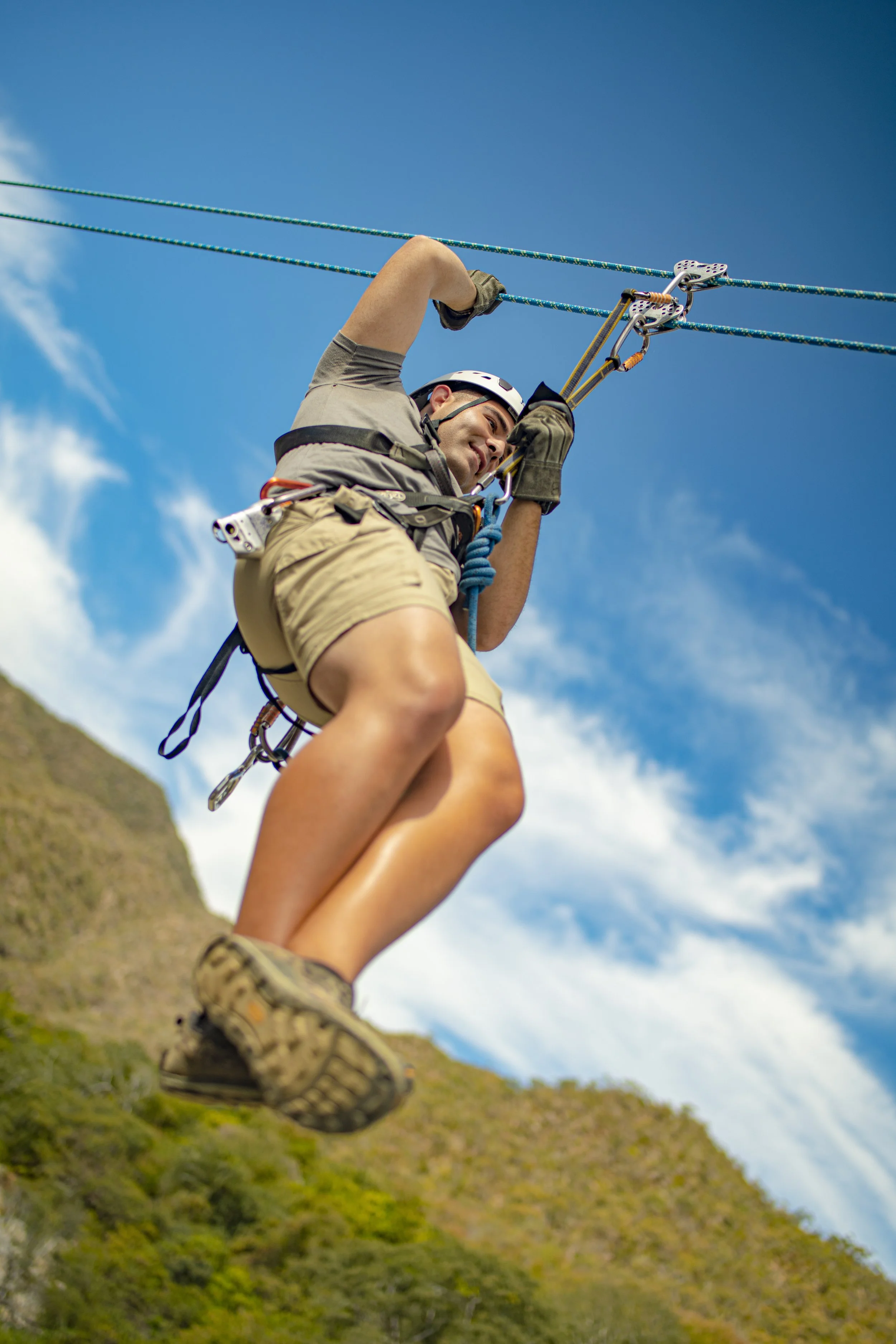 A person is ziplining across a landscape with green hillside and blue sky, wearing a helmet, gloves, and harness.
