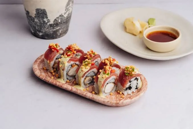 A plate of sushi rolls topped with fish, drizzled with sauce, and garnished with crunchy toppings, served with wasabi, ginger, and soy sauce, and a ceramic cup with a drink in the background.