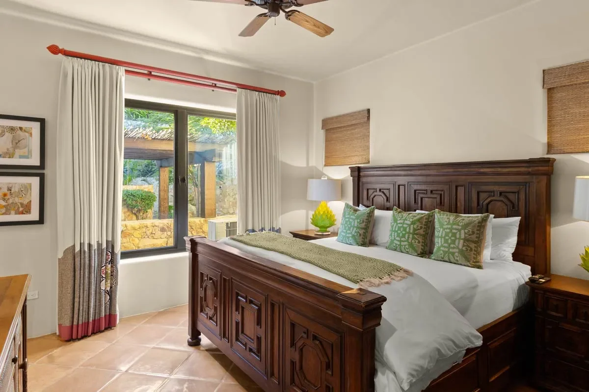 Hotel bedroom with wooden bed frame, white bedding, green pillows, nightstands with lamps, window with outdoor view and curtains, artwork on wall, ceiling fan.