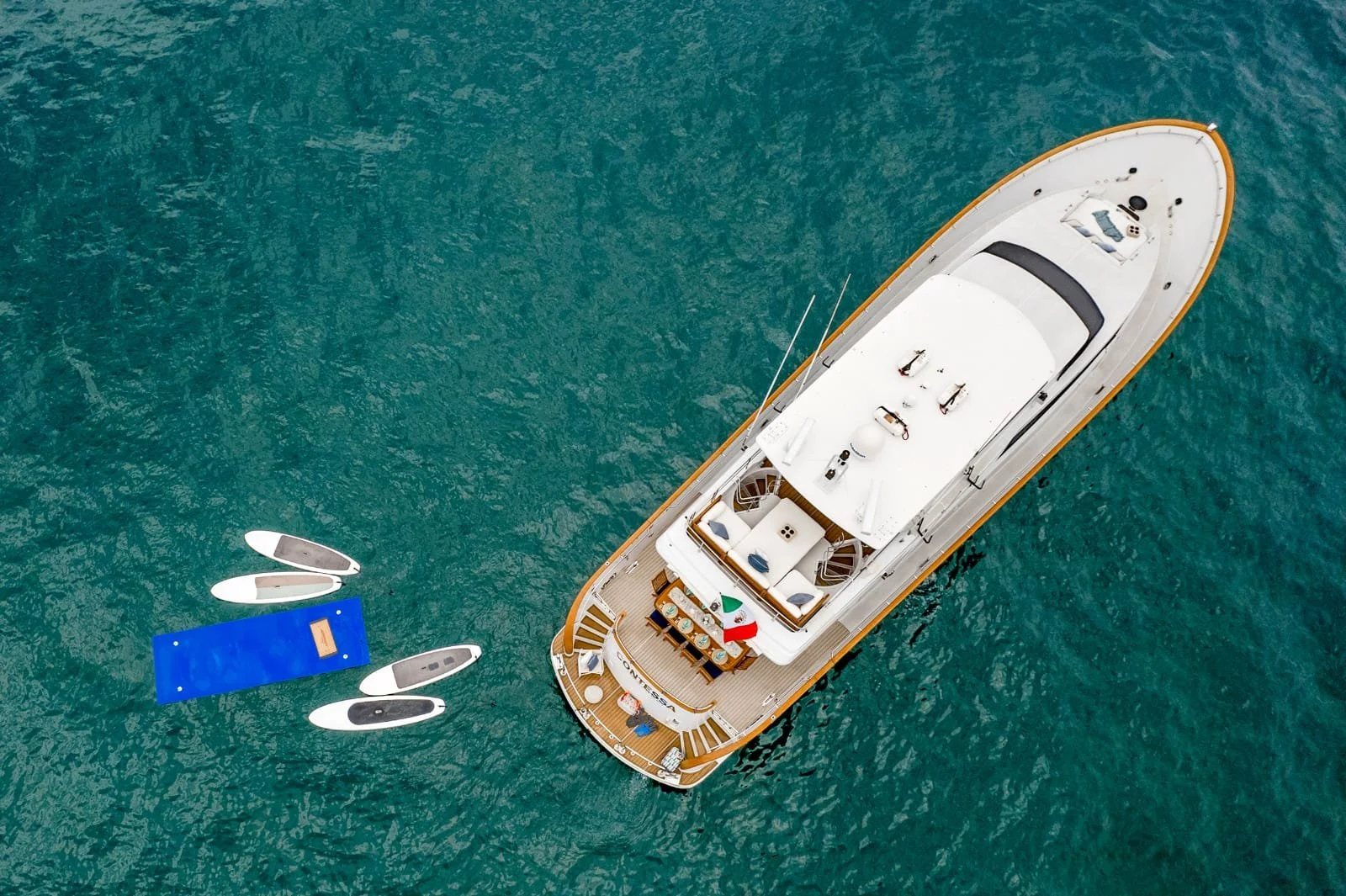 An aerial view of a large white yacht anchored in turquoise water, with a floating blue dock and four small boats nearby