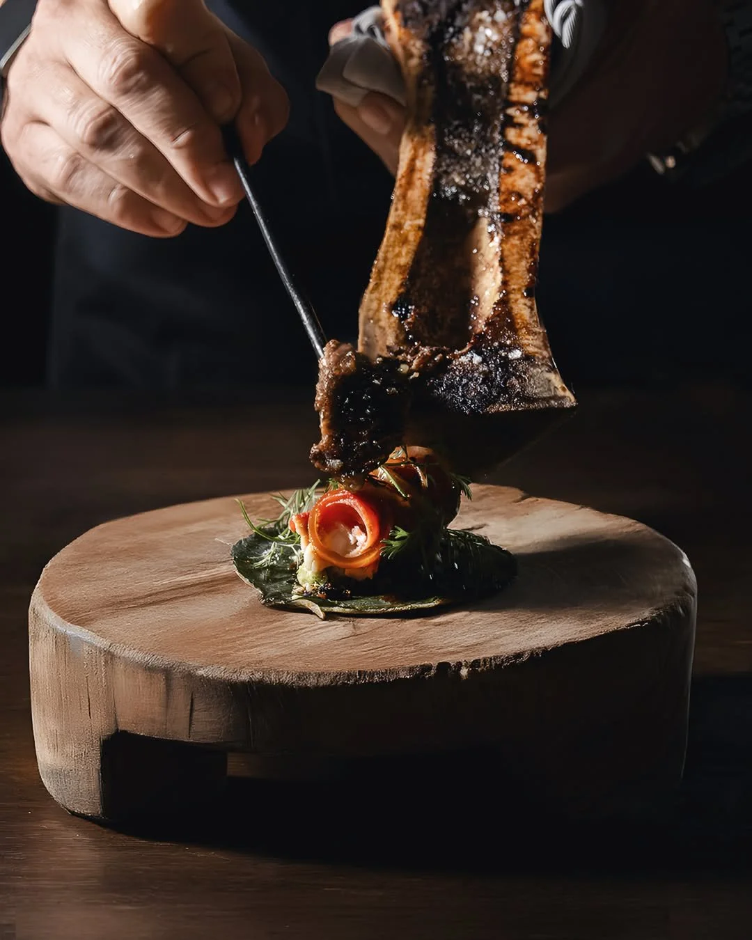 Chef garnishing a piece of grilled meat with herbs and tomato on a wooden serving board.